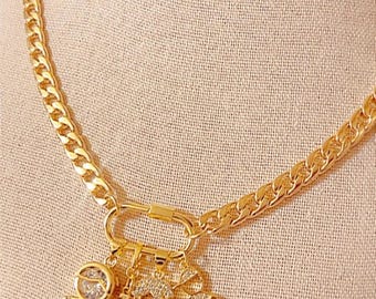 May include: Gold-colored necklace with a thick chain and a charm featuring three small figures and the word "Mama". The figures are embellished with small, clear stones. The necklace is displayed on a beige surface.
