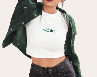 May include: White crop top with the word "sláinte" in green script. The top is paired with dark denim jeans and a dark green corduroy jacket. The outfit is casual and stylish.