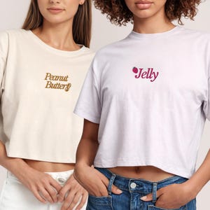 May include: Two women wearing white crop tops with different text. The top on the left says "Peanut Butter" in brown text. The top on the right says "Jelly" in pink text with a small red strawberry graphic.