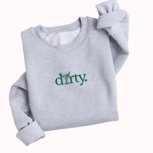 May include: A grey sweatshirt with a green embroidered design that says "dirty." The design includes a martini glass with a green olive.