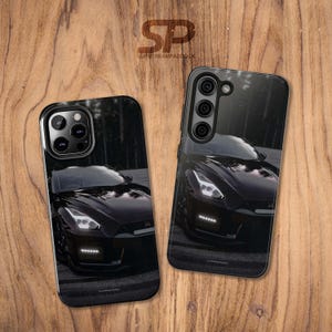 May include: Two black phone cases with a car design. The cases feature a dark image of a black sports car with bright headlights. The cases are on a wooden surface. The text "SLIPSTREAMPADDOCK" is visible.