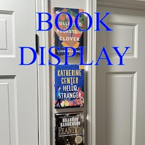 May include: A white door with three books displayed on a floating shelf. The books are "The Collected Regrets of Clover" by Alikk Brame, "Katherine Center Hello Stranger" and "Elantris" by Brandon Sanderson.
