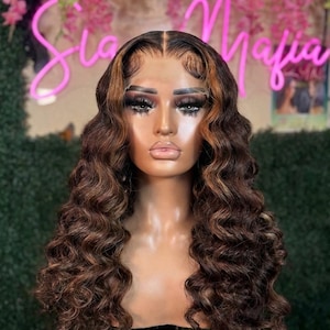 May include: A brown wig with long, wavy curls and highlights. The wig is styled on a mannequin head with a realistic face and makeup. The hair has a natural look with a dark brown base and lighter brown highlights. The wig is a fashion accessory.
