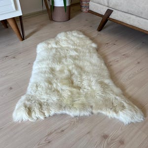 Puede incluir: Una alfombra de piel de oveja blanca con una textura suave y esponjosa. La alfombra está colocada sobre un suelo de madera clara.