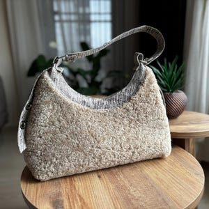 May include: A beige, textured shoulder bag with a patterned strap and trim. The bag is made of a soft, plush material and features a curved design. It sits on a wooden table, showcasing its shape and texture.