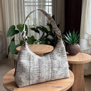 May include: A pale, textured shoulder bag with a curved design and a single strap. The bag is placed on a small wooden table. The background features a sofa, plants, and a window.