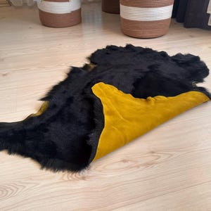 May include: A black and gold cowhide rug with a natural, irregular shape. The rug features a thick, shaggy black fur on one side and a smooth, mustard yellow suede on the other. The rug is displayed on a light wood floor.