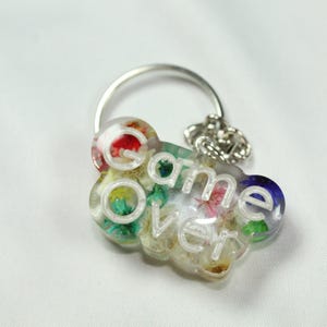 May include: A clear resin keychain shaped like a cloud with the text game over embossed in white. The interior of the resin contains small, colorful dried flowers in shades of red, yellow, green, and blue, attached to a silver metal ring.