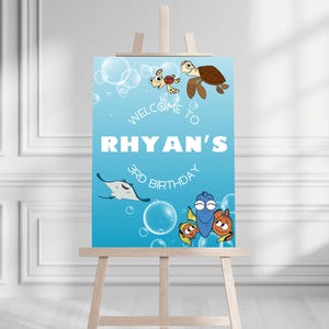 Puede incluir: Un cartel de bienvenida azul y blanco con el texto "Welcome to Rhyan's 3rd Birthday" e imágenes de dibujos animados de Dory, Nemo y una tortuga. El cartel está en un caballete de madera.