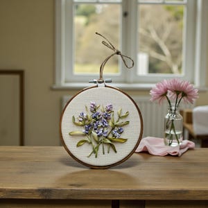 May include: A round embroidery hoop featuring a lavender floral design. The artwork showcases purple and green ribbon flowers on a cream-colored fabric, set within a wooden hoop. A twine loop is attached for hanging.