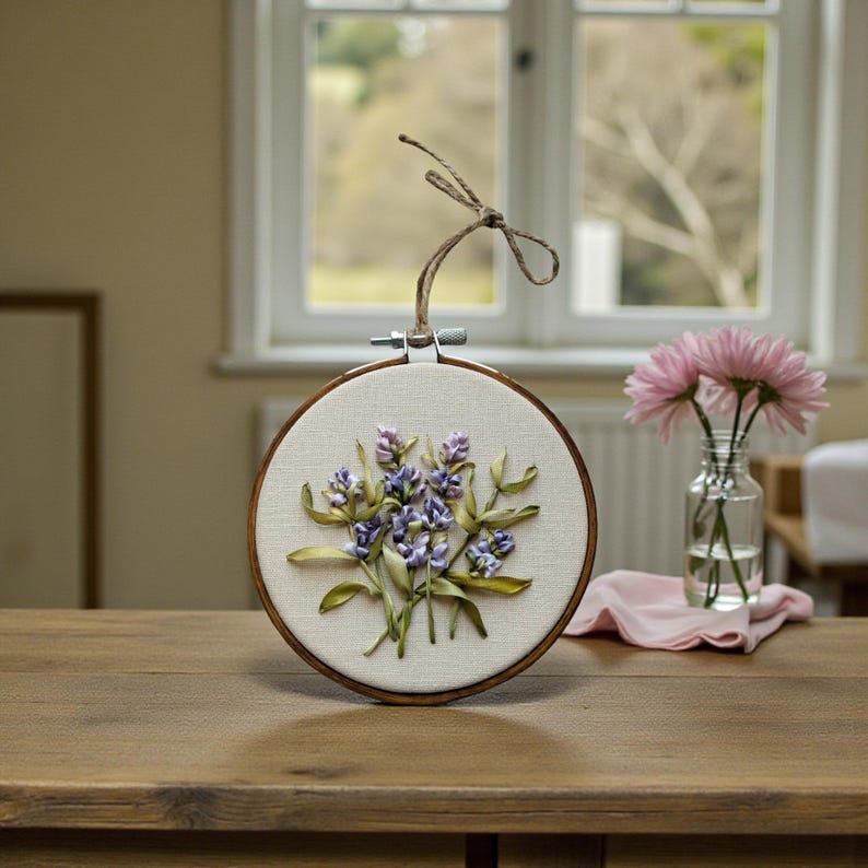 May include: A round embroidery hoop featuring a lavender floral design. The artwork showcases purple and green ribbon flowers on a cream-colored fabric, set within a wooden hoop. A twine loop is attached for hanging.
