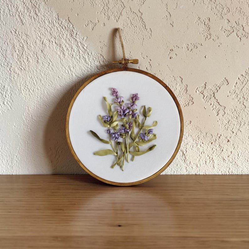 May include: A round, wooden embroidery hoop featuring a white fabric with a lavender floral design. The lavender is made of purple and green ribbon, and the hoop is suspended by a natural twine loop.
