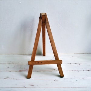 May include: A small, wooden easel with a natural finish. The easel has three legs and a horizontal bar at the bottom with the word "ART" printed on it. The easel is designed to hold a small canvas or artwork.