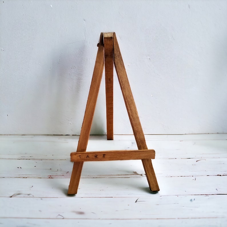 May include: A small, wooden easel with a natural finish. The easel has three legs and a horizontal bar at the bottom with the word "ART" printed on it. The easel is designed to hold a small canvas or artwork.