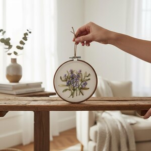 May include: A round embroidery hoop featuring a floral design with lavender-colored flowers and green leaves. The artwork is set against a cream-colored fabric and framed by a wooden hoop. A brown string is attached for hanging.