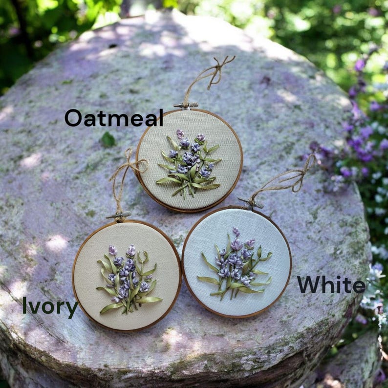 May include: Three round embroidery hoops with floral designs. The hoops are labeled "Oatmeal", "Ivory", and "White". Each hoop features a lavender bouquet with green leaves. The embroidery is set against a neutral fabric background, and the hoops are trimmed with a natural-colored wood.