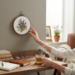 May include: A round, wooden embroidery hoop with a white fabric center, featuring a lavender floral design. The embroidery is mounted on a gray wall. A person's hand is touching the hoop. A cup of tea sits on a wooden table.