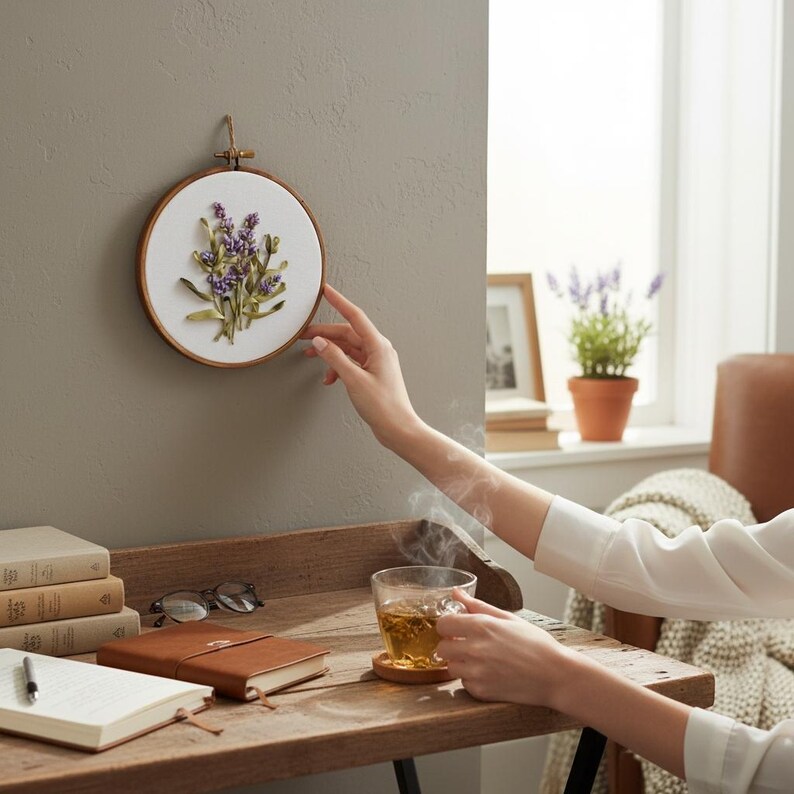May include: A round, wooden embroidery hoop with a white fabric center, featuring a lavender floral design. The embroidery is mounted on a gray wall. A person's hand is touching the hoop. A cup of tea sits on a wooden table.