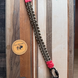 May include: A paracord lanyard featuring a braided olive drab cord with bright neon pink heat-shrink tubing at both ends. The lanyard is equipped with two black metal carabiners, one at each end, providing a secure attachment point for gear.