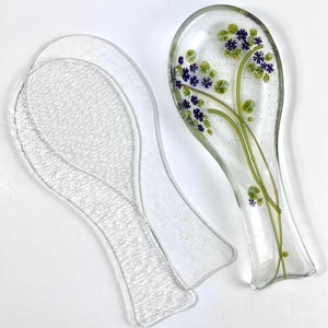 May include: Two clear glass spoon rests, one with a floral design featuring purple and green flowers. The spoon rests are textured and have a smooth, rounded shape.