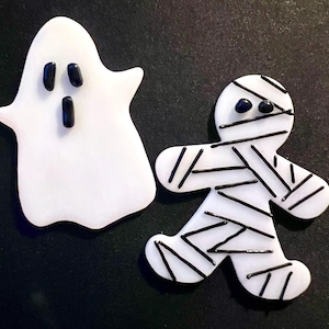 May include: Two white ceramic Halloween ornaments, one shaped like a ghost with black eyes and mouth, and the other shaped like a mummy with black eyes and white bandages.