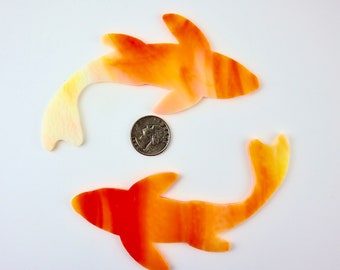 May include: Two orange and yellow glass fish shapes with a white background. The fish are about the size of a US quarter.