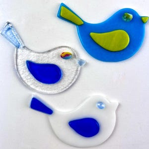 May include: Three colorful glass bird ornaments. One is blue with a yellow wing, another is clear with a blue wing, and the third is white with a blue wing. Each bird has a small, colorful eye and a simple, rounded shape.