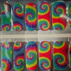 May include: A sheet of nail wraps featuring a vibrant tie-dye design. The wraps showcase a spiral pattern in shades of red, yellow, green, and blue. The nail wraps are in a silver package, with a hand holding the package.