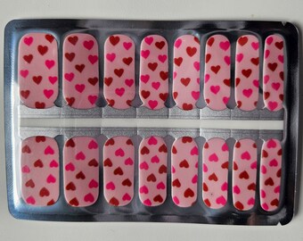 May include: A sheet of pink and red nail wraps with a heart pattern. The nail wraps are packaged in a clear plastic sheet with a silver backing.