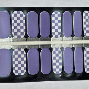 May include: A sheet of nail polish strips in a black package. The strips alternate between solid lavender and a white and lavender checkerboard pattern. The nail strips are in various sizes and shapes.