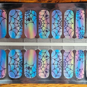 May include: A sheet of nail wraps with a Halloween theme. Each wrap features a black spider web design with a small spider. The background has a colorful, iridescent gradient of blues, pinks, and yellows. The wraps are in a clear plastic package.