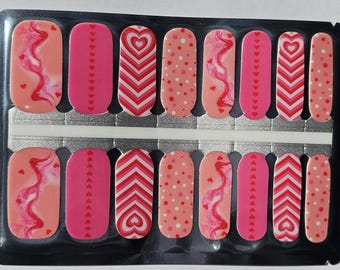 May include: A sheet of nail wraps with various designs in shades of pink and red. Patterns include hearts, swirls, dots, and chevron stripes. The nail wraps are packaged in a clear plastic container.