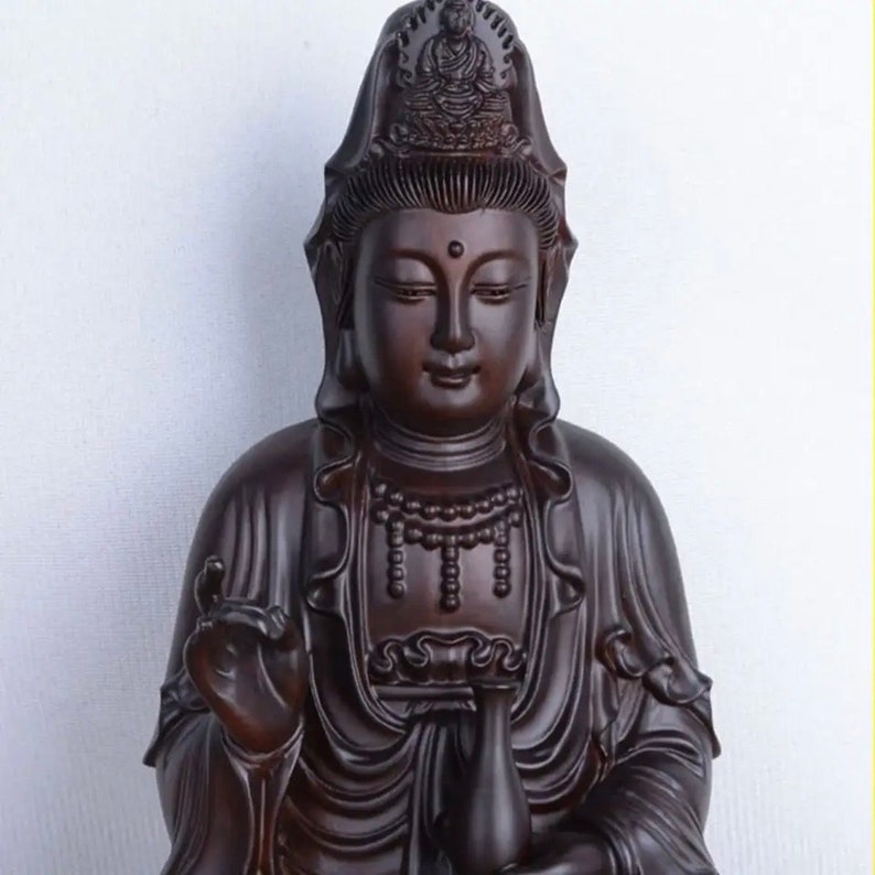 May include: A dark brown wooden statue of a serene figure with a detailed headdress and flowing robes. The statue features a delicate necklace and a gentle expression, with hands in a symbolic gesture. The background is a plain white.
