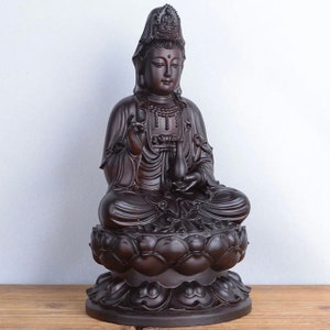 May include: A dark brown statue of a seated figure, possibly a deity, with serene facial features and ornate headdress. The figure is seated on a lotus flower base, holding a small vessel. The statue is likely made of wood or a similar material.
