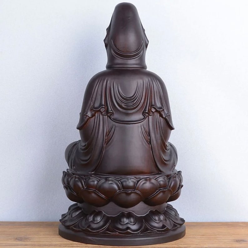 May include: A dark brown statue of a seated figure, viewed from the back. The figure is draped in flowing robes and sits atop a lotus flower base. The statue has a smooth, polished finish and is set against a light background.