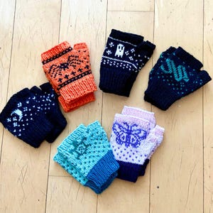 Puede incluir: Una colección de guantes sin dedos de punto en varios colores, incluyendo naranja, azul marino, turquesa y azul claro. Cada par presenta patrones únicos como una araña, un fantasma o una mariposa. Los guantes están colocados sobre una superficie de madera.