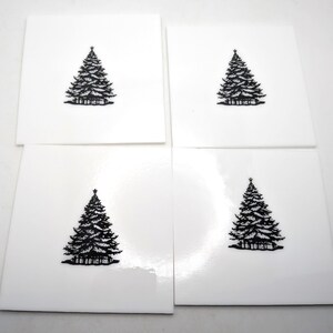 May include: A set of four square white coasters, each featuring a centered black graphic illustration of a Christmas tree. The coasters have a smooth, glossy finish and a clean, minimalist design, displaying a consistent tree motif on every piece.