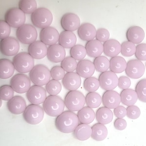 May include: A collection of light pink, round, cabochon-shaped glass beads of varying sizes. The beads are arranged in a scattered pattern, with a glossy finish. These beads are suitable for jewelry making and craft projects.