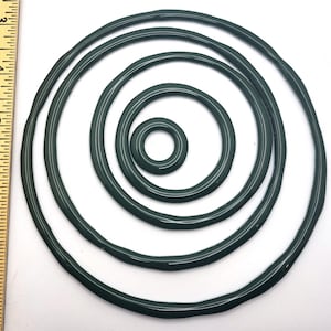 May include: A collection of dark green rubber O-rings of varying sizes, arranged in a nested circular pattern. The rings have a smooth, glossy finish and a white stripe detail. A ruler is visible on the left side of the image, providing scale.