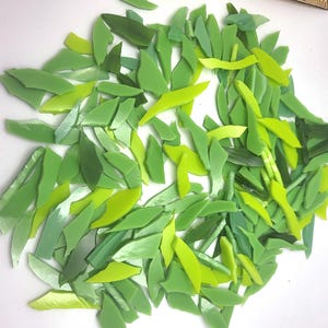 May include: A collection of irregularly shaped glass pieces in various shades of green, including light, medium, and dark tones. The pieces are scattered, suggesting they are for mosaic or craft projects. The glass has a glossy finish.