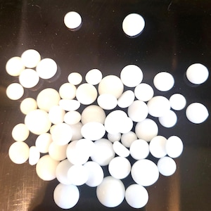 May include: A collection of white, spherical objects of varying sizes, scattered on a dark surface. The objects appear to be smooth and opaque, with a consistent white color. The arrangement is random, with some objects overlapping.