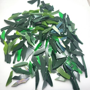 May include: A collection of irregularly shaped glass pieces in various shades of green, from light to dark, scattered on a white surface. The pieces have a reflective, glossy finish, suggesting they are for mosaic or craft projects.