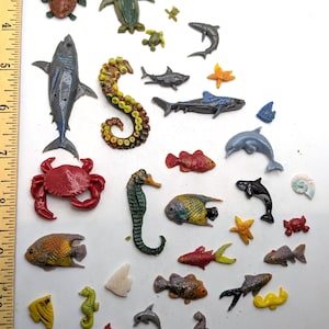 May include: A collection of miniature plastic sea creatures including sharks, fish, crabs, seahorses, and dolphins, arranged next to a yellow ruler showing measurements in inches. The figures are various colors and sizes, depicting marine life.