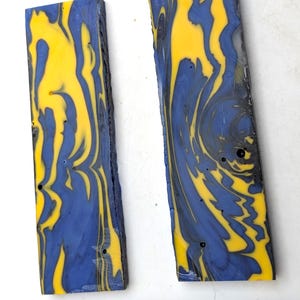 May include: Two rectangular blocks of resin featuring a marbled pattern of vibrant yellow and deep blue swirls. The blocks are positioned above a wooden ruler that shows measurements in inches, indicating the pieces are approximately 3 inches long.