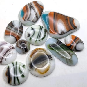 May include: A collection of ten smooth, polished glass cabochons in various shapes and sizes. Each piece features a white base with swirling patterns of orange, brown, black, green, and blue, creating a marbled effect across the surfaces.