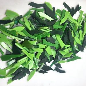 May include: A collection of irregularly shaped glass pieces in various shades of green, from light to dark. The pieces are scattered across a white surface, suggesting they are for a mosaic or craft project.