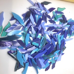 May include: A collection of irregularly shaped glass pieces in shades of blue, teal, and purple. The pieces vary in size and shape, creating a mosaic effect. The glass has a glossy finish, reflecting light.
