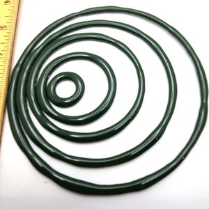 May include: A collection of dark green rubber O-rings of varying sizes, arranged in a spiral pattern. The rings are circular and appear to be made of a flexible material. A ruler is visible on the left side of the image, providing a size reference.