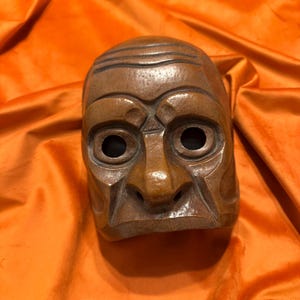 May include: A wooden mask with a carved face, featuring deep-set eyes, a prominent nose, and a wide mouth. The mask is painted in shades of brown and has a textured surface.