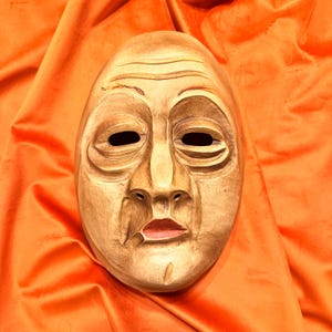 May include: A wooden mask with a carved face, featuring deep-set eyes, a prominent nose, and a slightly downturned mouth. The mask is painted in shades of brown and beige, with a hint of pink on the lips.